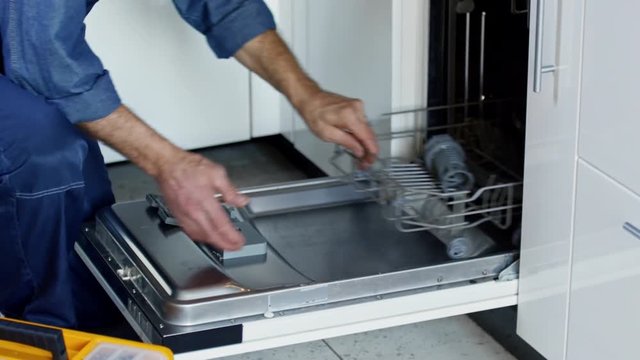 Tilt down of mature male worker in overalls finishing installation of new dishwasher machine in clients kitchen