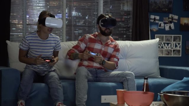 Young couple wearing VR headsets and playing with gamepads while having fun on couch at home