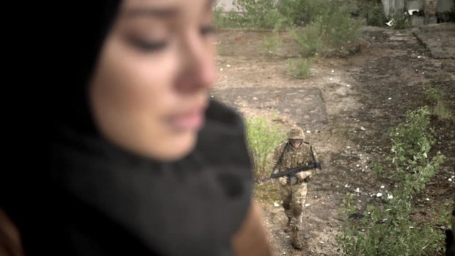 Young sad muslim woman in hijab turning and looking at camera, armed man with weapon walking outside, rainy day, terrorism concept