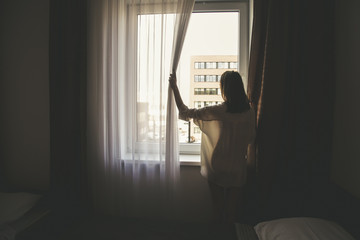 Photo of beautiful woman opening curtains and looking through the window