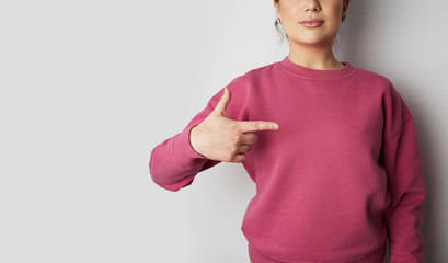 Young hipster girl wearing pink hoody and pointing finger with space for your logo or design. Mock-up of pink hoody on white empty wall in the background. Copy paste text space