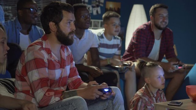 Group Of Multiethnic Men And Teens Gathering On Sofa At Home And Playing Videogame With Gamepads