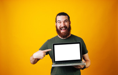 Happy bearded hipster pointing at his blank white screen of the notebook on yellow background.