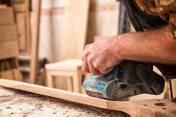 Young man builder carpenter equals polishes wooden board with a  random orbit sander  in the...