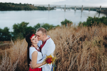 A man kisses a woman in the cheek, a woman smiles happily. The couple stands embracing in the high dry grass by the river. Ukrainian patriots. Ukrainian ethnic wedding on Khortytsya.