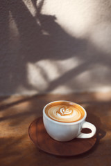 Cup of cappuccino with beautiful heart shape latte art. Tropical and hipster vibes. Plants shadows. Sul lights and shadows.