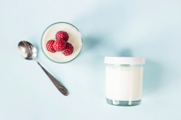 Open jar with homemade yogurt and raspberries next to a closed jar on a blue background. Top view, flat lay