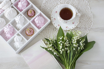 on the table a cup of tea, a book, a bouquet of lilies of the valley, a box of marshmallows, meringues and cakes