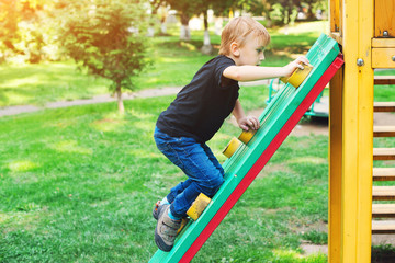 Fototapeta na wymiar Happy kid climbing on outdoor playground. Little blond boy having fun outdoors. Summer, spring and autumn leisure for active kids. Child climbs confidently up on kids playground. Summer holidays.