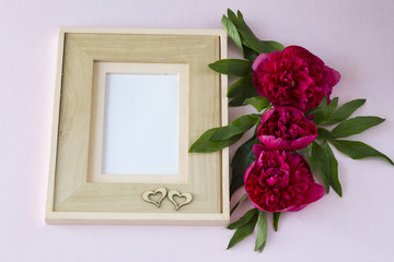 on a pink background, three pink peonies, an empty frame for a photo and two wooden hearts