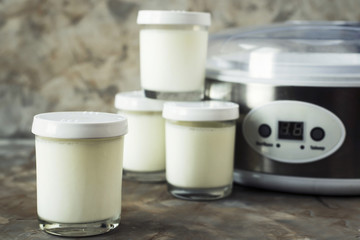 Four jars with homemade yogurt next to a yoghurt machine on a gray background. Dietary food