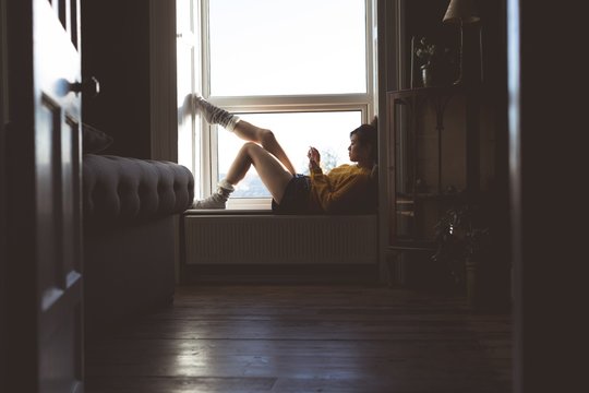 Woman using smartphone while relaxing on widow sill at home