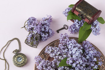 on a pink background a lilac bouquet in an old bronze plate, a bronze small cage, a pocket old watch, an old chest and a key