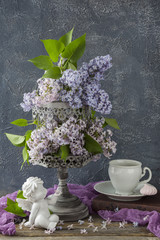 on the wooden table an old gray cage and in it a bouquet of lilac, a book, a cup, a meringue and a statue of a white angel