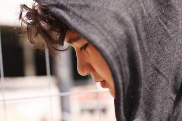 Sad troubled teenager school boy with hood on posing outdoor sitting alone on the street - stock...