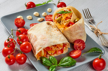 Shawarma sandwich with fresh vegetables and sauce on the gray plate decotated with cherry tomatoes, basil leaves, knife and fork. Lavash snack - traditional arabic food. Eastern cuisine. Close up view