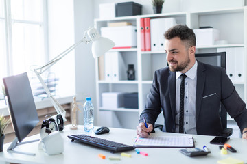 A man sits in the office at the table, holds a pen and works with documents and a computer.