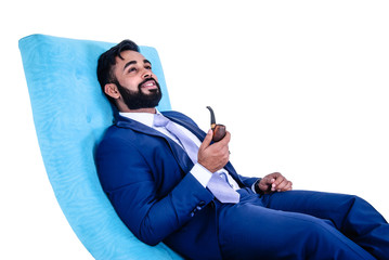 Handsome young bearded Asian man wearing blue suit sitting in sofa with a pipe, isolated on white background