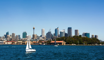 Fototapeta na wymiar Sailboat crossing the deep blue waters of Sydney Harbor against a backdrop of the city skyline