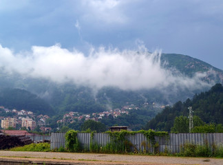 Landscape: a white cloud descended on the village at the foot of a forested mountain. The gray sky is above this city, soon will be rain