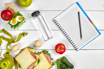 Top view planning notebook with copy space and healthy breakfast. Sandwich with ham and cheese, eggs, cucumber, tomato and bottle of water on white wooden table.