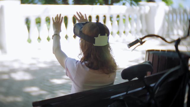 Young woman uses head-mounted display in the park. Playing game using VR-helmet for smart phones. Happy