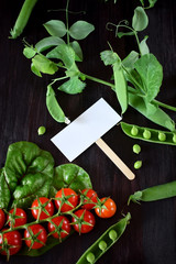Cherry tomatoes, chard and green peas are framing an empty card in the middle against the dark background. Copy space