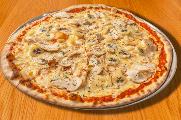 large round pizza with unusual toppings of chicken, banana, pinapple and cream, with the normal tomato and cheese on a metal dish on a wooden table
