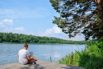 man working on laptop while sitting on wooden dock. legs in river water