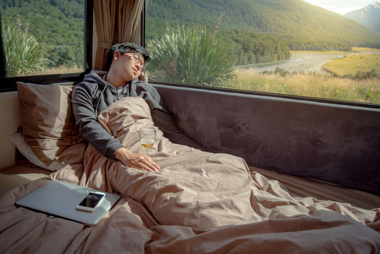 Young Asian man hangover after drinking beer in camper van with mountain scenic view through the window, laptop computer and smartphone on the bed. digital nomad on road trip concept