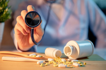 Male Asian doctor holding stethoscope in hospital clinic. Pills spilling out of pill bottles on doctor desk. Medical treatment and health care consultation concepts.
