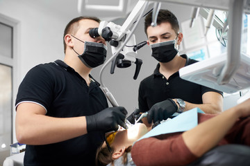 Male dentist in a black uniform fixing patient's teeth while another one is helping him with...