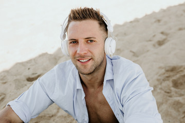 Portrait of Young hipster man in a white shirt and jeans listening to music in headphones on a smartphone and is sitting on beach.Digital Music lounge and relaxing concept. Cropped
