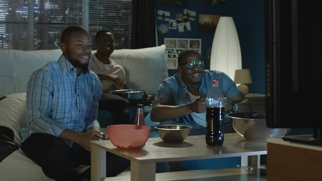 Group of young African-American men chilling on sofa with popcorn and having fun while playing videogame with gamepad