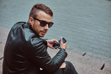 Fashionable guy dressed in a black jacket and jeans holds a smartphone sitting on steps.