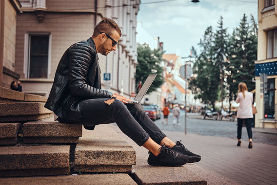 Fashionable freelancer guy dressed in a black jacket and jeans working on the laptop while sitting on steps against an old building in Europe.