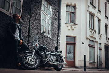 Naklejka na ściany i meble Fashionable biker dressed in a black leather jacket and jeans leaning on a wall near his retro motorcycle on old Europe street.