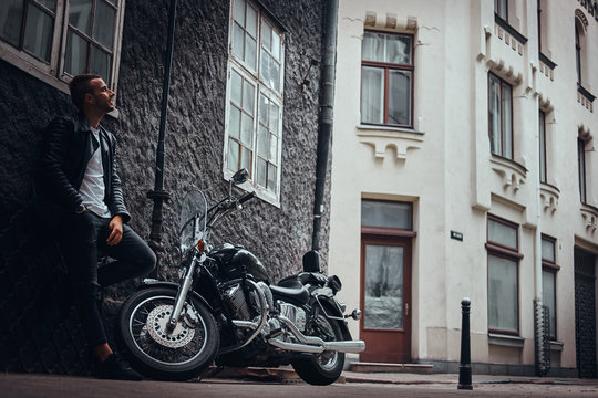 Fashionable biker dressed in a black leather jacket and jeans leaning on a wall near his retro motorcycle on old Europe street.