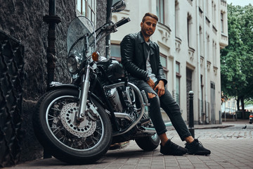 Fototapeta na wymiar Fashionable biker dressed in a black leather jacket and jeans sitting on his retro motorcycle on an old Europe street.