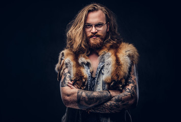 Tattoed redhead hipster male with long luxuriant hair and full beard dressed in a t-shirt and jacket holds the fox skin on a shoulder in a studio. Isolated on the dark background.