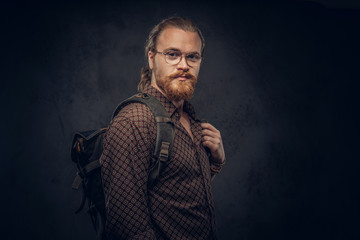 Portrait of a redhead hipster student in glasses dressed in a brown shirt, holds a backpack, posing at a studio. Isolated on the dark background.