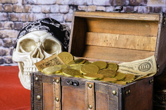 human skull with black skull cap next to wood chest full of gold coins and old paper money on red with brick wall background