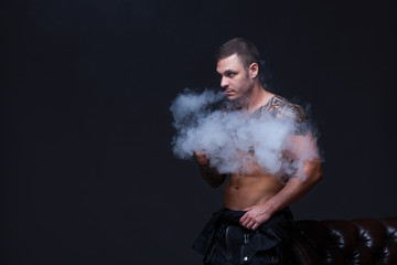 Vaper. The man with a muscular naked torso with tattoos smoke an electronic cigarette on the dark background