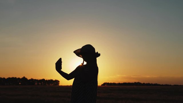 The young girl makes selfie in a hat at sunset, the dress develops in the wind. slow motion, silhouette.