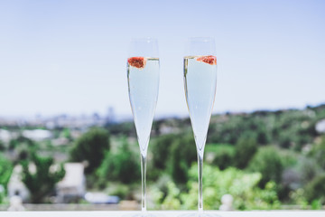 Two champagne glasses with strawberry on sunny terrace outdoor patio overlooking clean blue sky at summer day outside of the city