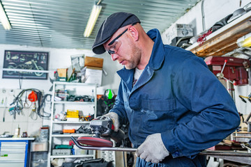 A young man welder in a blue T-shirt, goggles and construction gloves processes metal an angle grinder in the garage, in the background a lot of tools