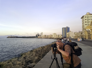 Photographer with camera during sunset with Atlantic Ocean, residential building and Morro Castle in background - Malecon, Havana, Cuba