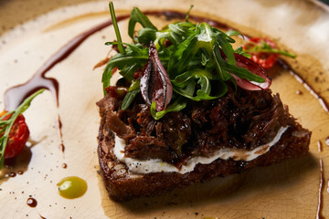 Italian Bruschetta with lamb beef. Open sandwich with Dark rye bread, dried cherry tomatoes, rosemary, olives and arugula on plate at luxury restaurant table, top view. Delicious snack food