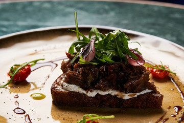 Italian Bruschetta with lamb beef. Open sandwich with Dark rye bread, dried cherry tomatoes, rosemary, olives and arugula on plate at luxury restaurant table, top view. Delicious snack food