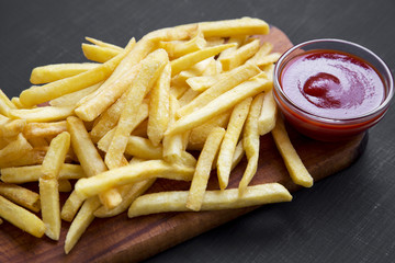 Fried tasty french fries with ketchup on wooden board over black background, top view.
